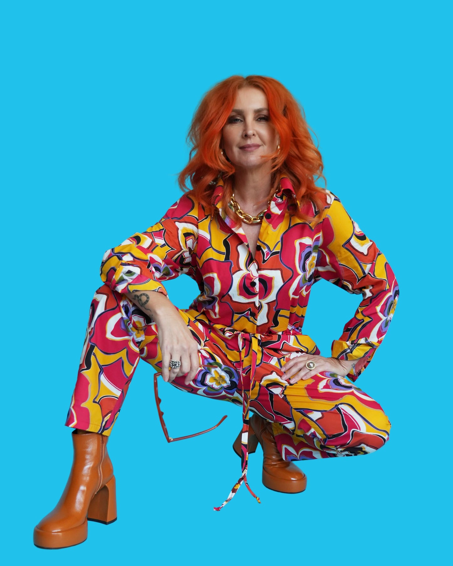 A person wearing a cherry red boiler suit with a vibrant palm Royale floral pattern, featuring a button-through front fastening, shirt collar, and cuffed sleeves. The suit includes a waistband with a drawstring and side pocket detail, designed to be versatile and comfortable for all-season wear.