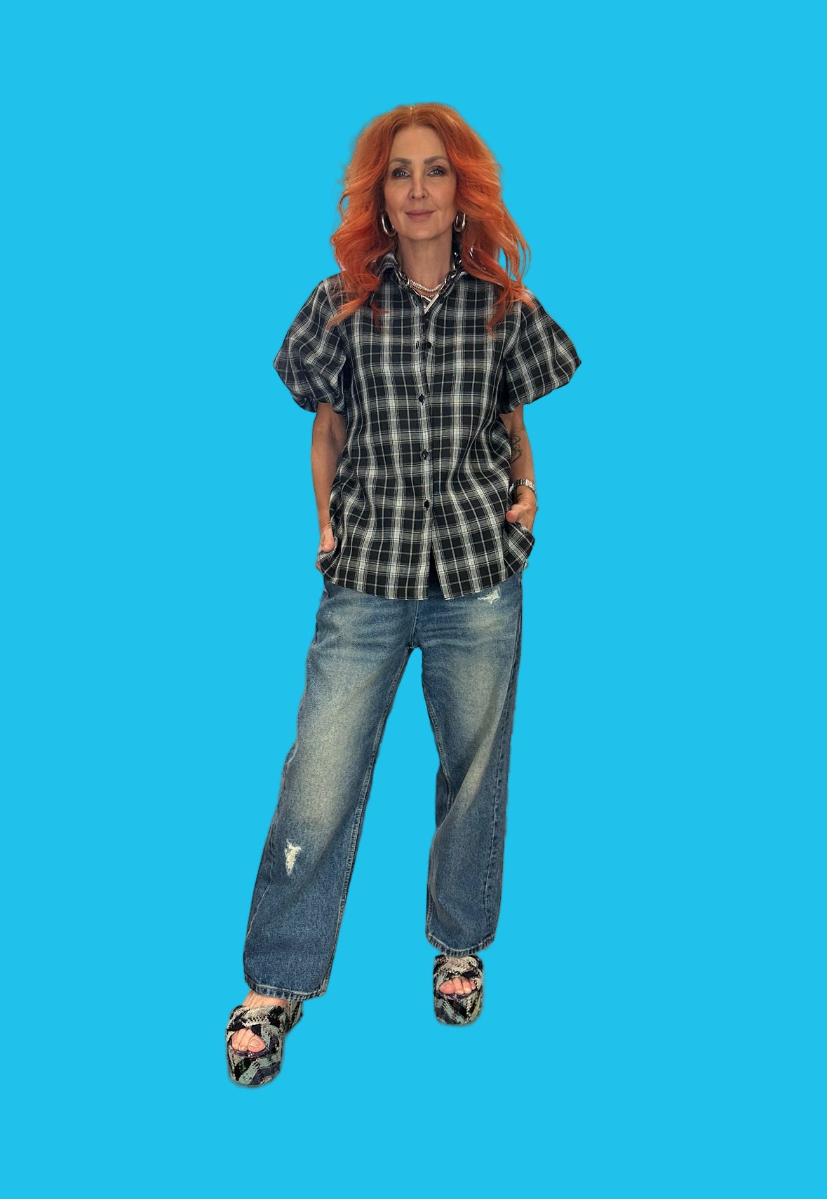 Woman with red hair wearing a plaid shirt and jeans against a blue background